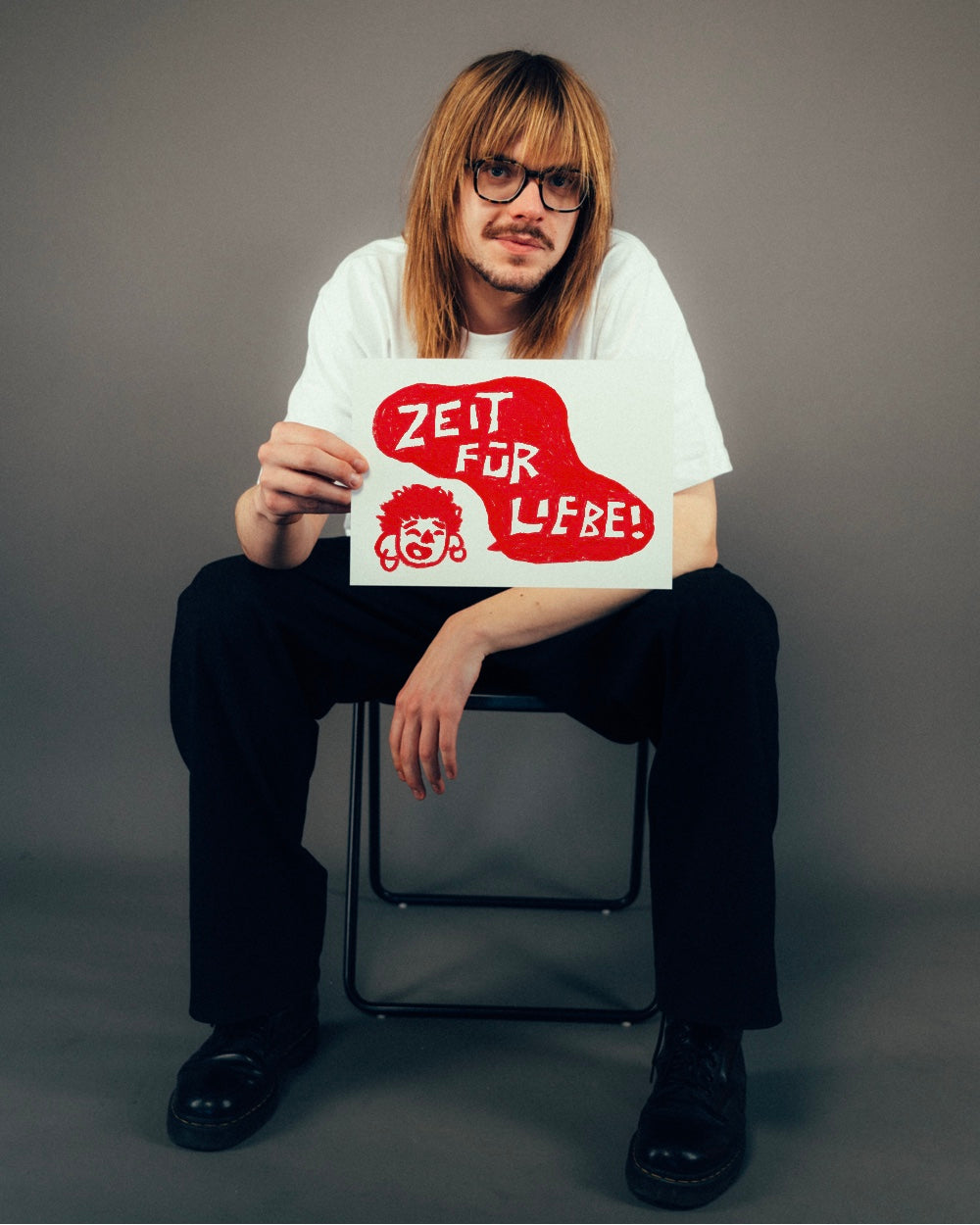 Person sitzt auf einem Stuhl in neutralem Studiohintergrund und hält ein Blatt mit handgezeichneter Illustration hoch. In kräftigem Rot steht in einer organisch geformten Sprechblase der Schriftzug „ZEIT FÜR LIEBE!“, daneben ein lachendes Gesicht mit Ohrringen und lockigem Haar. Der Druck auf dem weißen T-Shirt ist dezent sichtbar – im Fokus steht das farbintensive, verspielte Motiv mit positiver Botschaft.