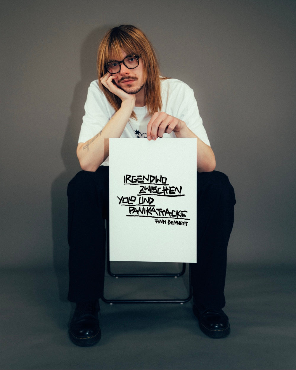 Person sitzt auf einem Stuhl in neutraler Studio-Umgebung und hält ein Blatt mit handschriftlichem Spruch in schwarzer Markeroptik. Darauf steht: „IRGENDWO ZWISCHEN YOLO UND PANIKATTACKE“ – mit dem Namenszusatz „FINN BENNETT“. Die Schrift ist betont, leicht schräg und unterstrichen. Der Fokus liegt auf dem humorvoll-nachdenklichen Printmotiv im minimalistischen Stil.