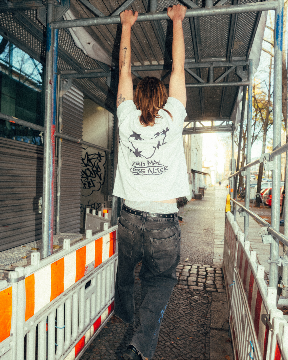 Rückansicht einer Person, die ein weißes T-Shirt mit schwarzem Rückendruck trägt. Das Motiv zeigt mehrere Dornenherzen, die kreisförmig angeordnet sind, und darunter den Schriftzug „ZEIG MAL LIEBE ALTER“. Das Shirt wird mit einer dunklen Jeans und einem Nietengürtel kombiniert. Die Person hängt mit ausgestreckten Armen an einem Baugerüst in einer urbanen Straßenszene.