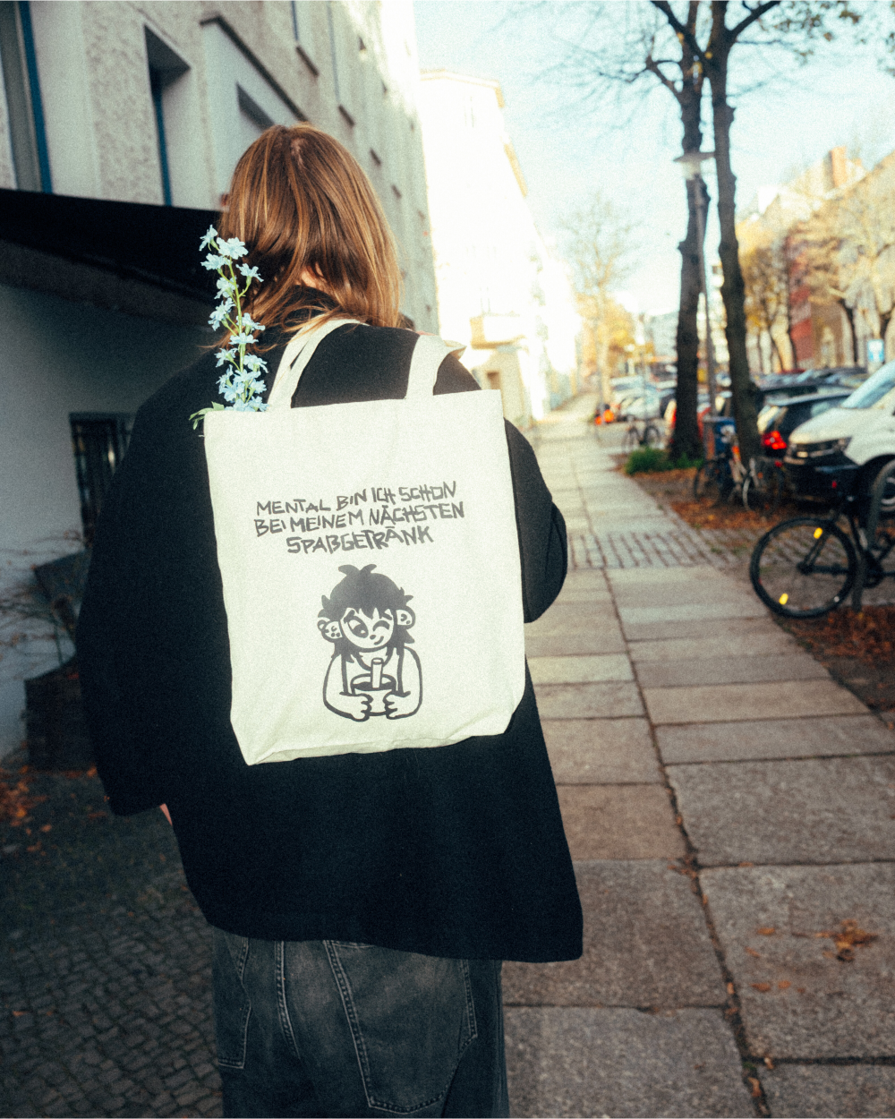 Person geht auf einem Bürgersteig entlang und trägt eine beigefarbene Stofftasche mit schwarzem Print auf dem Rücken. Das Motiv zeigt eine gezeichnete Figur mit Becher und den Schriftzug „MENTAL BIN ICH SCHON BEI MEINEM NÄCHSTEN SPASSGETRÄNK“. Aus der Tasche ragen hellblaue Blumen hervor.