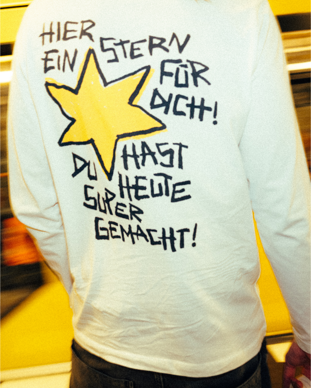 Nahaufnahme eines weißen Longsleeves mit großem Rückendruck. Das Design zeigt einen gelben, handgezeichneten Stern, daneben und darunter steht in schwarzer, verspielter Schrift: „HIER EIN STERN FÜR DICH! DU HAST HEUTE SUPER GEMACHT!“. Der Druck wirkt wie mit Filzstift gestaltet, aufgetragen auf weichem Baumwollstoff. Im Hintergrund ist eine gelbe U-Bahn in Bewegung erkennbar.