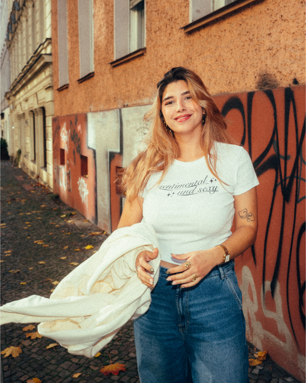Weiße, figurbetonte Crop-T-Shirt mit schwarzem Schriftzug „sentimental and sexy“ auf der Brust, gestaltet in geschwungener Schreibschrift mit kleinen Sternsymbolen. Die Person trägt dazu eine blaue Jeans und goldenen Schmuck und hält eine helle Jacke in der Hand. Aufgenommen in einer herbstlichen Straßenszene mit Graffitiwand und Laub am Boden.