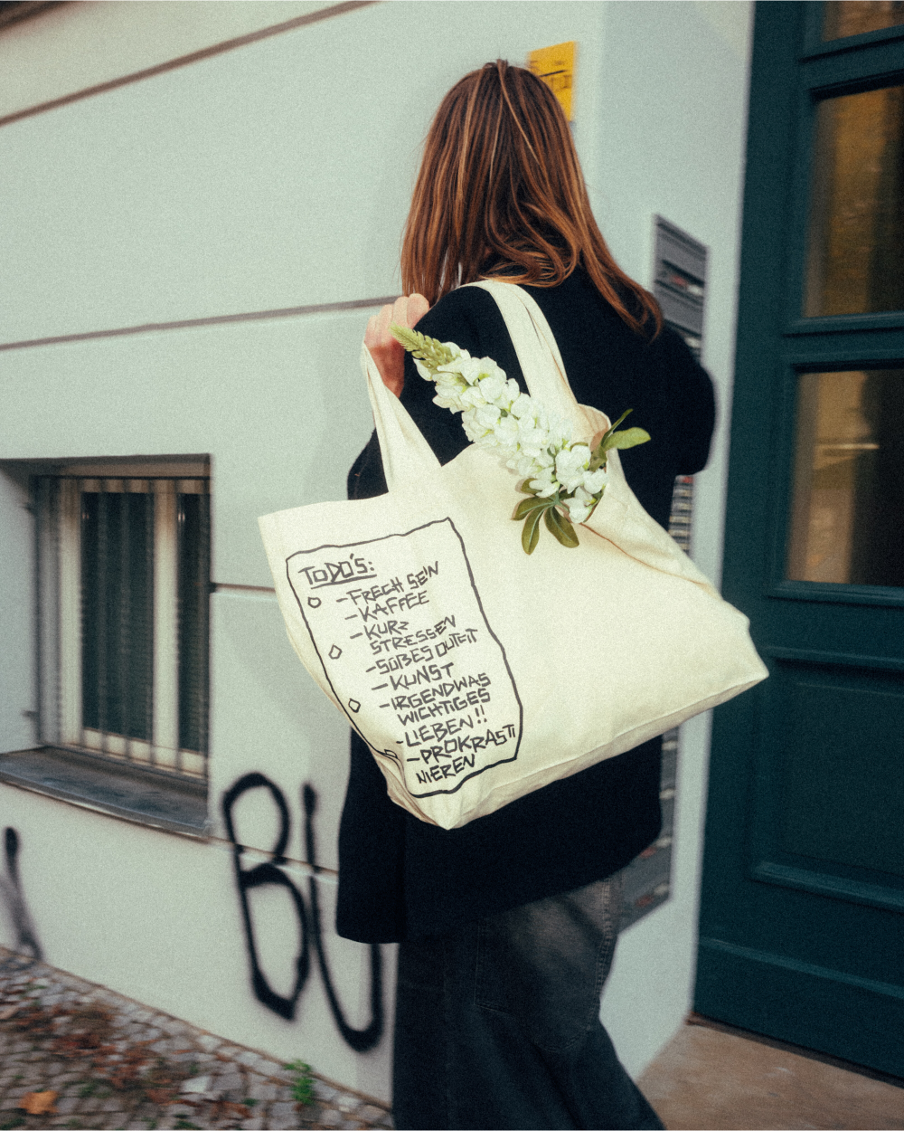 Beiger Stoff-Shopper aus fester Baumwolle mit schwarzem, handgezeichnetem To-do-Listen-Print auf der Vorderseite. Auf dem Print stehen humorvolle Punkte wie „FRECH SEIN“, „KAFFEE“, „KURZ STRESSEN“, „SÜßES OUTFIT“, „KUNST“, „IRGENDWAS WICHTIGES“, „LIEBEN!!“ und „PROKRASTINIEREN“. Die Tasche wird über der Schulter getragen und enthält weiße Blumen – lässiges Alltagsaccessoire mit verspieltem Illustrationsdesign.