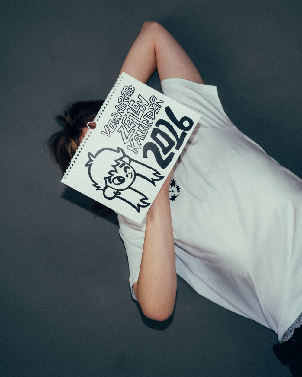 Person liegt auf dem Rücken und hält einen aufgeklappten Spiral-Kalender vor das Gesicht. Auf der Kalenderseite steht handgezeichnet der Schriftzug „Verwirrte Zeilen Kalender 2026“ neben einer Illustration eines Gesichts mit langen Haaren. Der Kalender ist in Schwarz-Weiß gestaltet und zeigt einen verspielten, handgezeichneten Stil.