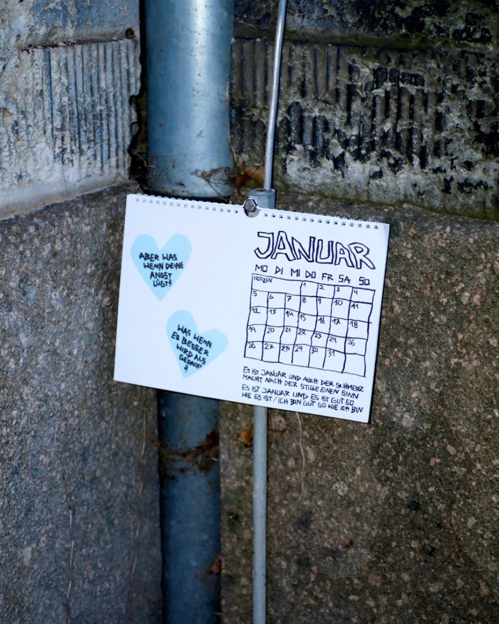 Kalenderblatt für den Monat Januar, aufgehängt an einer grauen Steinwand. Die Seite zeigt ein handgezeichnetes Monatsraster mit Datumsspalten und zwei blauen Herzen, in denen handschriftlich motivierende Sprüche stehen. Der Kalendertext unten ist ebenfalls handgeschrieben und im charakteristischen, illustrativen Stil gestaltet.
