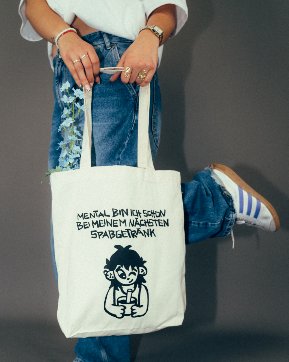 Person steht in legerer Pose und hält eine beigefarbene Stofftasche mit schwarzem Printmotiv. Die Tasche zeigt eine Illustration einer Figur mit Becher sowie den Schriftzug „MENTAL BIN ICH SCHON BEI MEINEM NÄCHSTEN SPASSGETRÄNK“. Aus der Tasche ragen hellblaue Blumen. Die Person trägt ein weißes T-Shirt, eine locker sitzende Jeans und helle Sneaker.