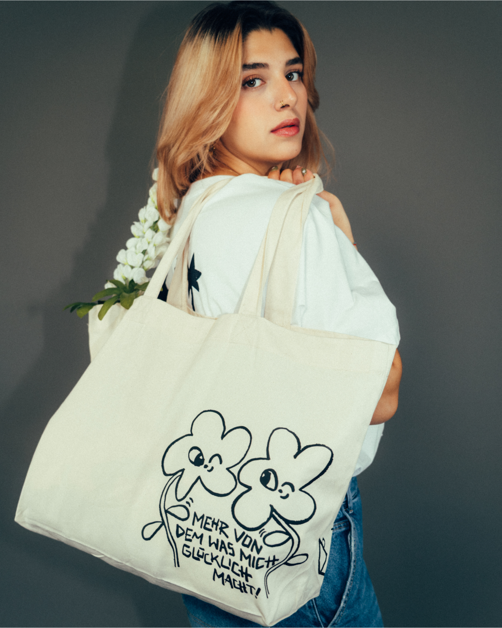 Beiger Baumwoll-Shopper mit schwarzem Print von zwei gezeichneten vierblättrigen Kleeblättern mit Gesichtern. Unter den Figuren steht der handgeschriebene Schriftzug „Mehr von dem, was mich glücklich macht!“. Die Tasche wird locker über der Schulter getragen, mit weißen Blüten befüllt. Lässige Stofftasche mit positiver Botschaft und verspieltem Design.