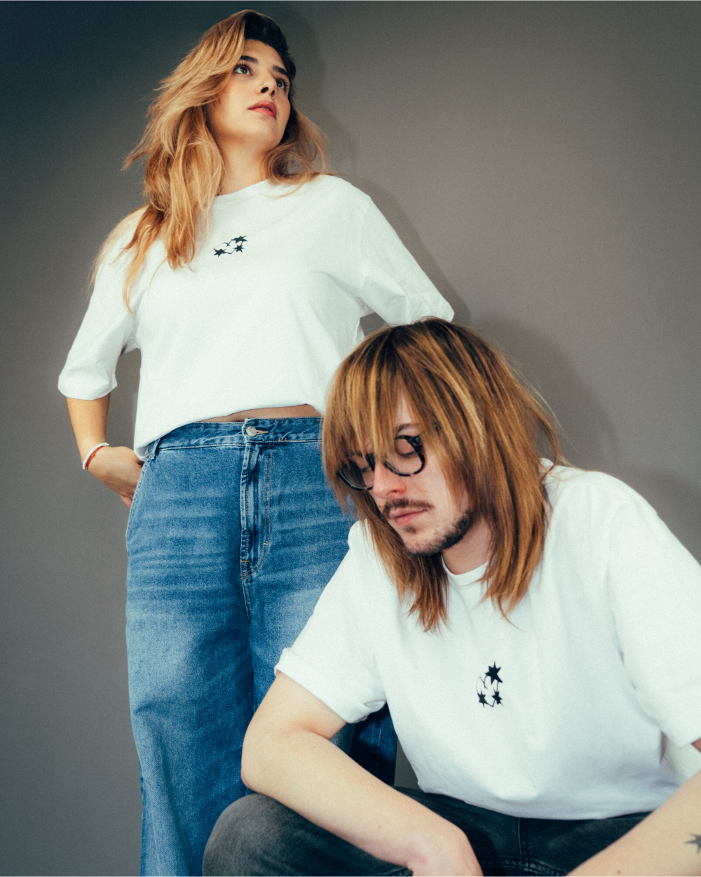 Zwei Personen tragen weiße T-Shirts mit kleinem schwarzen Frontprint auf der linken Brust. Das Motiv zeigt drei ineinander verschlungene Dornenherzen. Die stehende Person kombiniert das Shirt mit einer hellblauen Jeans mit hohem Bund, die sitzende Person mit einer dunklen Jeans. Das Foto ist in neutraler Studio-Umgebung aufgenommen und hebt die Frontansicht der Shirts hervor.