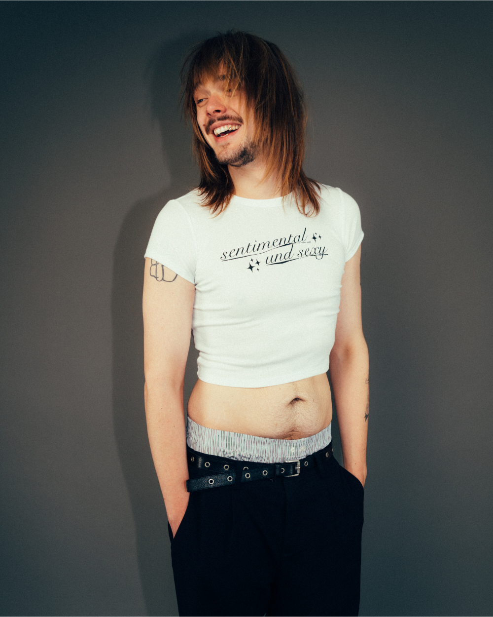 Weißes, eng geschnittenes Crop-T-Shirt mit schwarzem Schriftzug „sentimental and sexy“ auf der Brust in verspielter Schreibschrift mit kleinen Sternsymbolen. Das Shirt wird in Kombination mit einer schwarzen Hose und Gürtel getragen, vor neutralem grauem Hintergrund fotografiert.