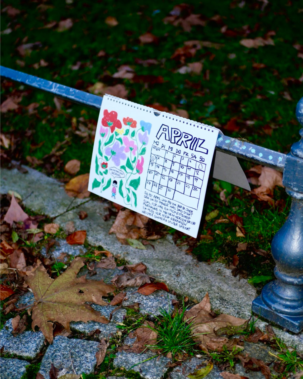 Kalenderseite für den Monat April, die an einem Metallgeländer im Freien lehnt. Links ist eine bunte handgezeichnete Blumenillustration mit einer kleinen Figur zu sehen, rechts ein Monatsraster mit dem Schriftzug „April“ in verspielter schwarzer Schrift. Darunter steht ein kurzer handgeschriebener Text. Die Szene ist von Herbstlaub und Kopfsteinpflaster umgeben.