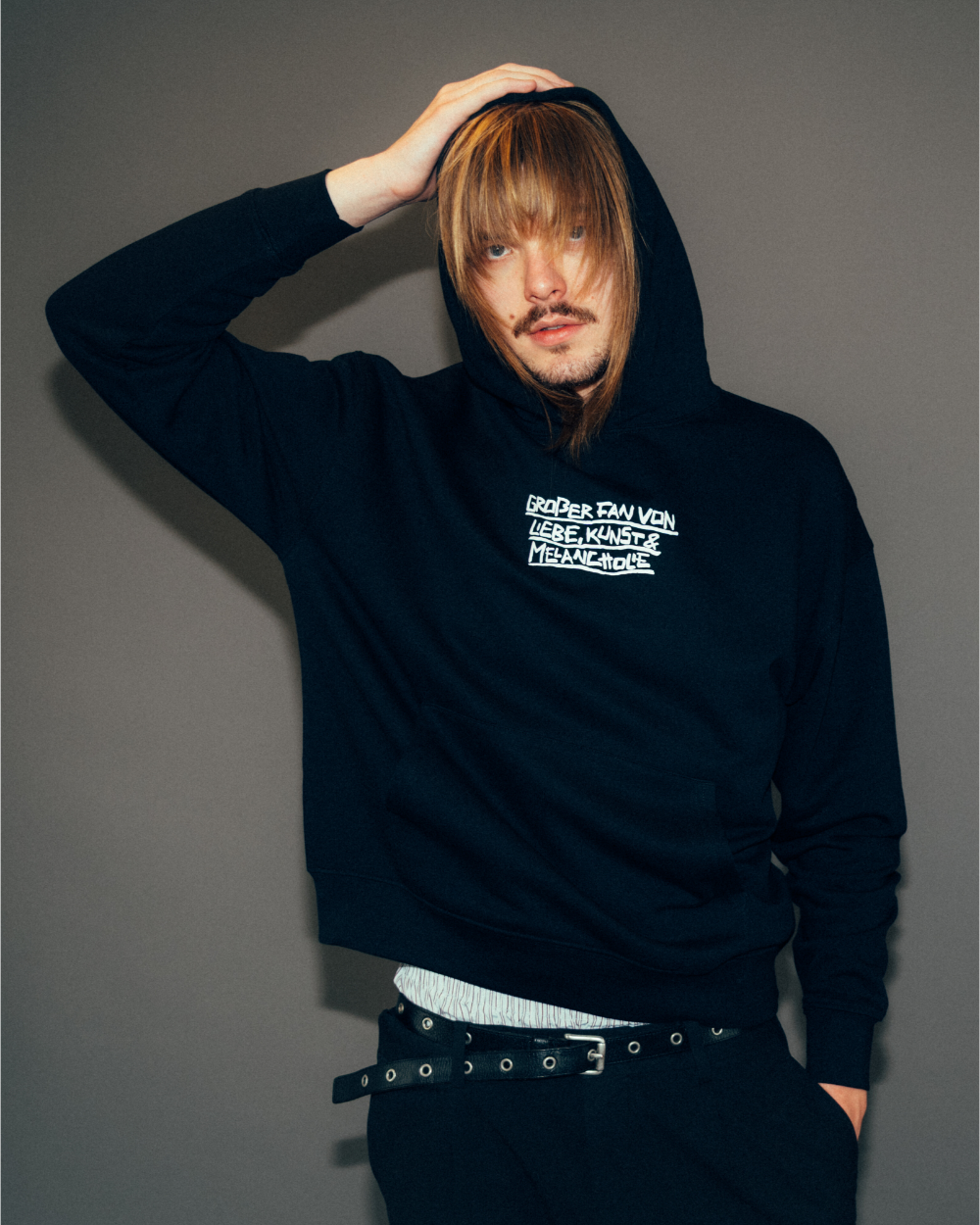Schwarzer Hoodie mit weißem Frontprint in handschriftlicher Optik. Der mittig platzierte Schriftzug lautet: „GRÖßER FAN VON LIEBE, KÜNSTEN & MELANCHOLIE“. Der Kapuzenpullover hat eine große Kängurutasche, eine doppellagige Kapuze ohne Kordelzug und gerippte Bündchen an Ärmeln und Saum.