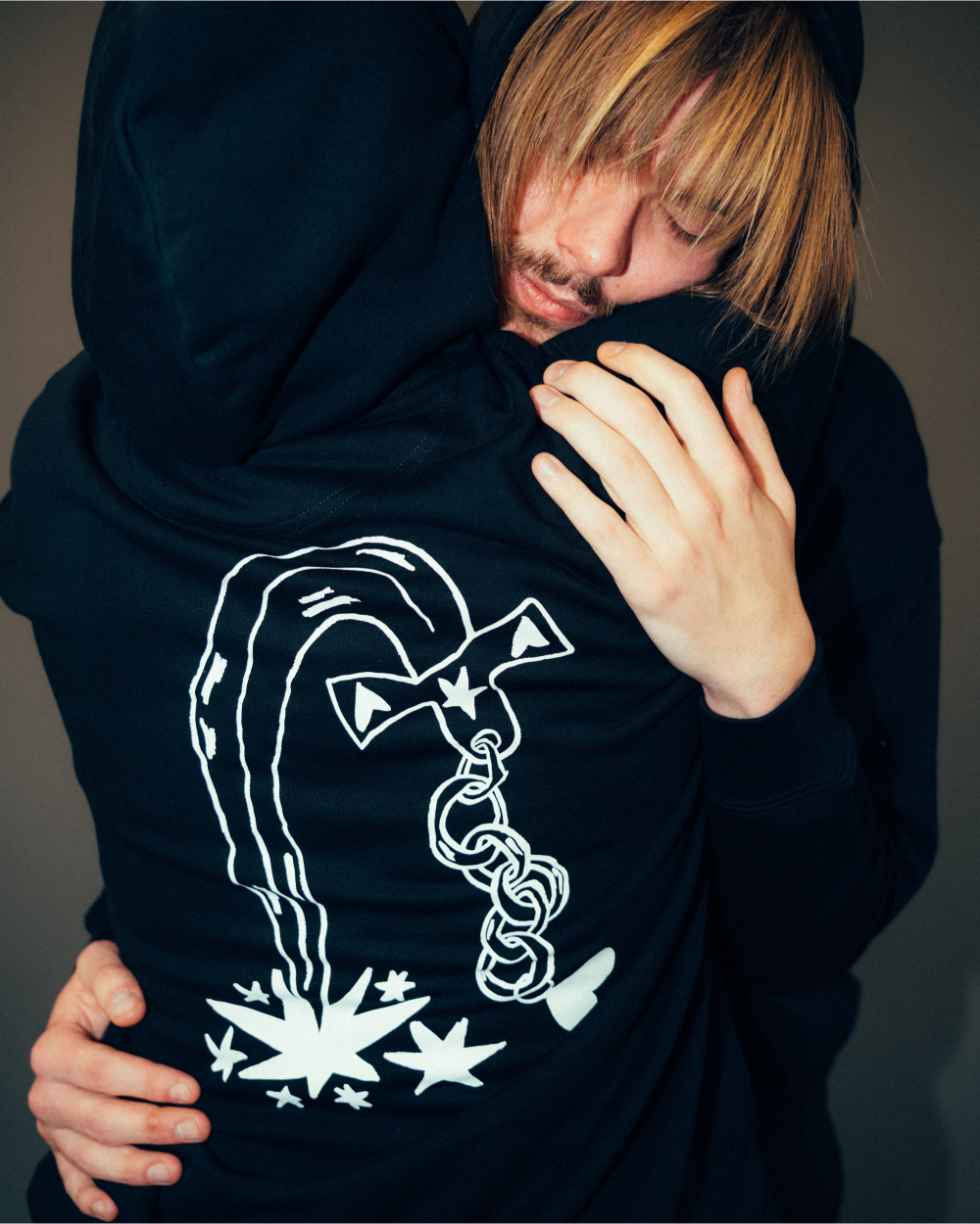 Schwarzer Kapuzenpullover mit großem weißen Rückendruck. Das Motiv zeigt eine stilisierte Kette mit Schleifen- und Sternsymbolen, aus der grafisch dargestellte Sterne und Linien hervorgehen. Der Hoodie besteht aus weicher Baumwollmischung, hat eine Kapuze ohne Kordelzug und eine lockere, gemütliche Passform.