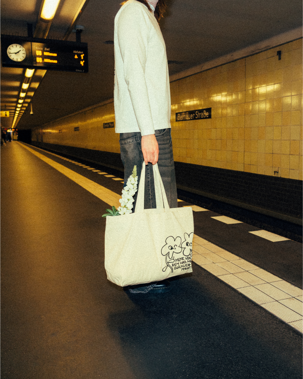 Beiger Stoff-Shopper aus Baumwolle mit schwarzem Print von zwei gezeichneten vierblättrigen Kleeblättern und dem handschriftlichen Schriftzug „Mehr von dem, was mich glücklich macht!“. Die Tasche wird locker in der Hand getragen und enthält weiße Blumen. Robuster, alltagstauglicher Shopper mit positiver Botschaft und verspieltem Illustrationsmotiv.