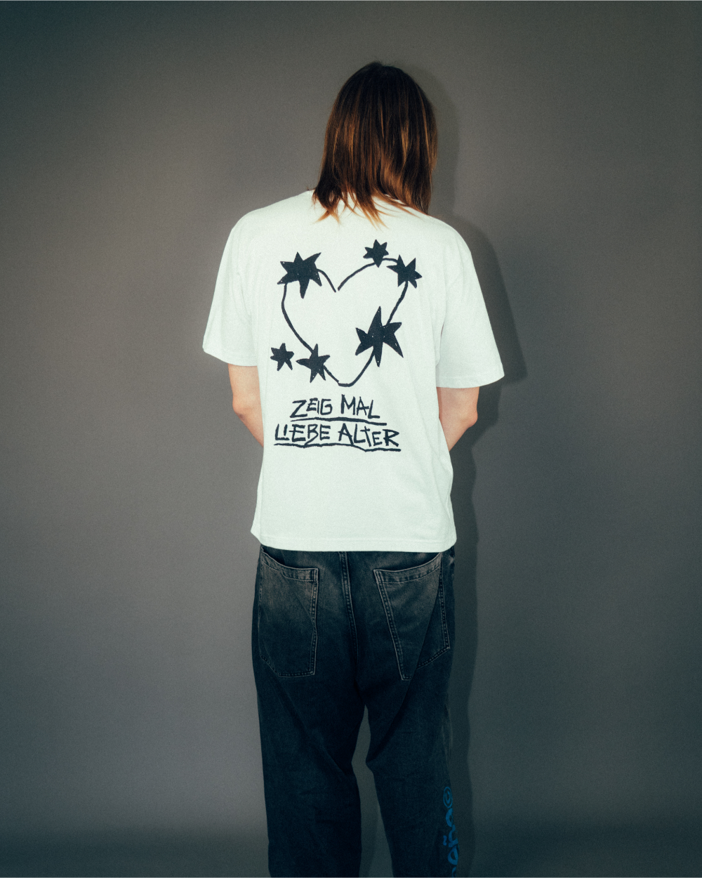 Rückansicht einer Person, die ein weißes T-Shirt mit großem schwarzen Rückendruck trägt. Das Motiv zeigt ein aus Dornen geformtes Herz mit mehreren sternförmigen Elementen sowie den darunter platzierten Schriftzug „ZEIG MAL LIEBE ALTER“. Das Shirt wird mit einer dunklen Jeans kombiniert und vor neutralem grauem Hintergrund präsentiert.