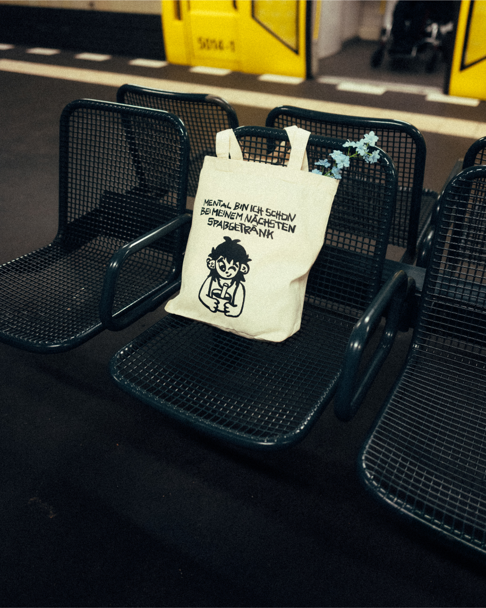Eine beigefarbene Stofftasche mit schwarzem Print liegt auf einem U-Bahn-Sitz aus dunkelgrünem Gittermetall. Der Aufdruck zeigt eine gezeichnete Figur mit Becher und den Schriftzug „MENTAL BIN ICH SCHON BEI MEINEM NÄCHSTEN SPASSGETRÄNK“. Aus der Tasche ragen hellblaue Blumen. Im Hintergrund ist eine gelbe U-Bahn zu sehen.