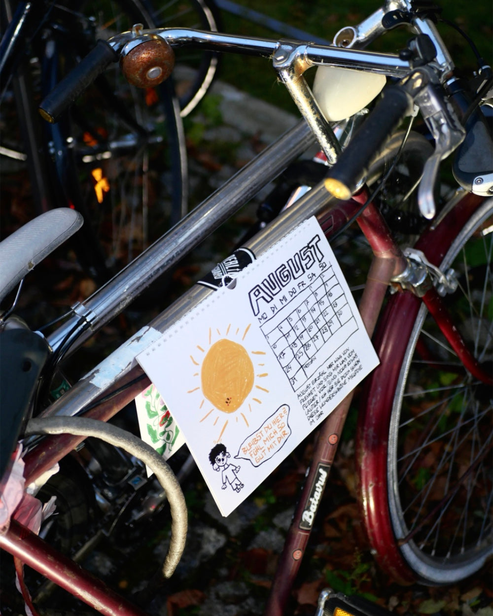 Kalenderseite für den Monat August, aufgehängt an einem Fahrradlenker. Das Blatt zeigt eine handgezeichnete Sonne mit Gesicht, eine kleine Figur und den Schriftzug „August“ über einem Monatsraster. Darunter steht ein humorvoller handgeschriebener Text. Die Illustration ist in Schwarz, Gelb und Orange gehalten und vermittelt sommerliche Stimmung.