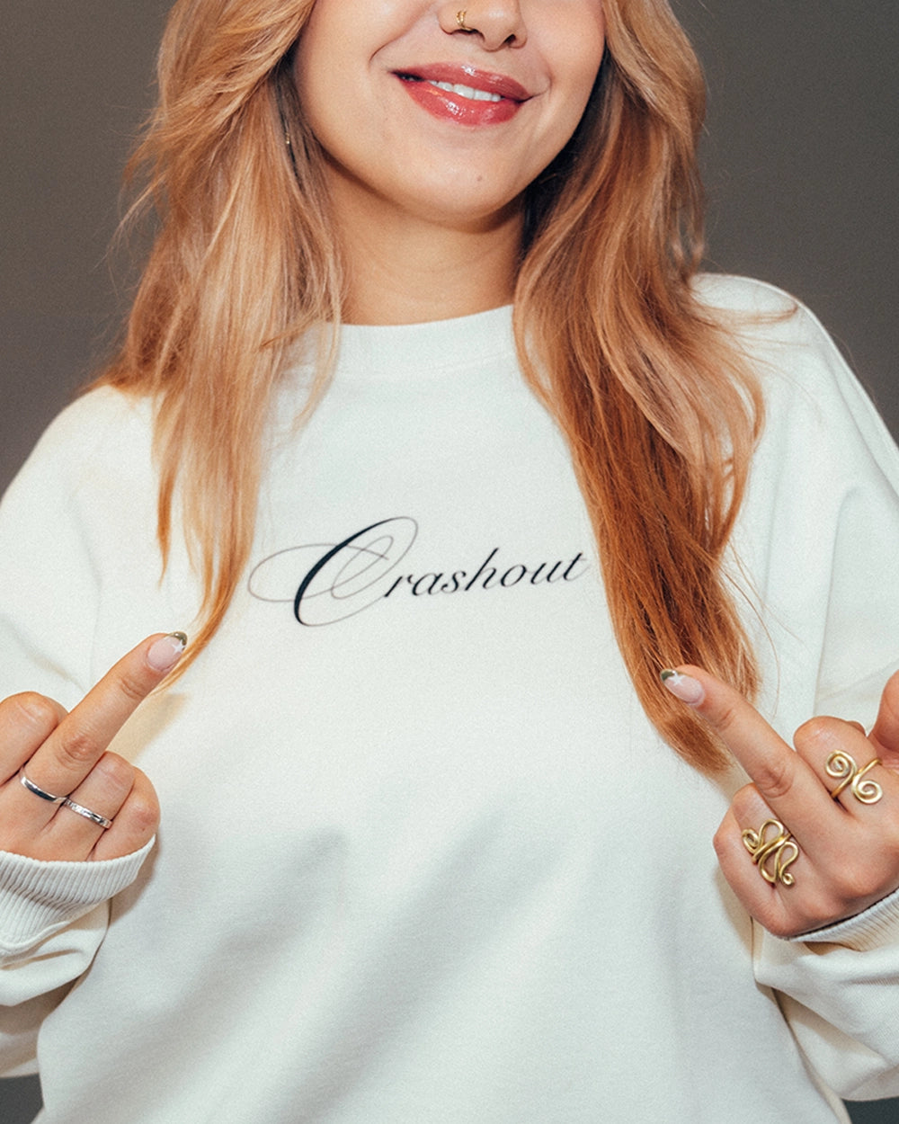 Eine Person trägt ein cremefarbenes Sweatshirt mit dem schwarzen Schriftzug „Crashout“ in eleganter Schreibschrift auf der Brust. Die Person zeigt beide Mittelfinger, trägt goldene und silberne Ringe und hat hellbraunes, offenes Haar. Der Fokus liegt auf dem Oberteil und dem Aufdruck.