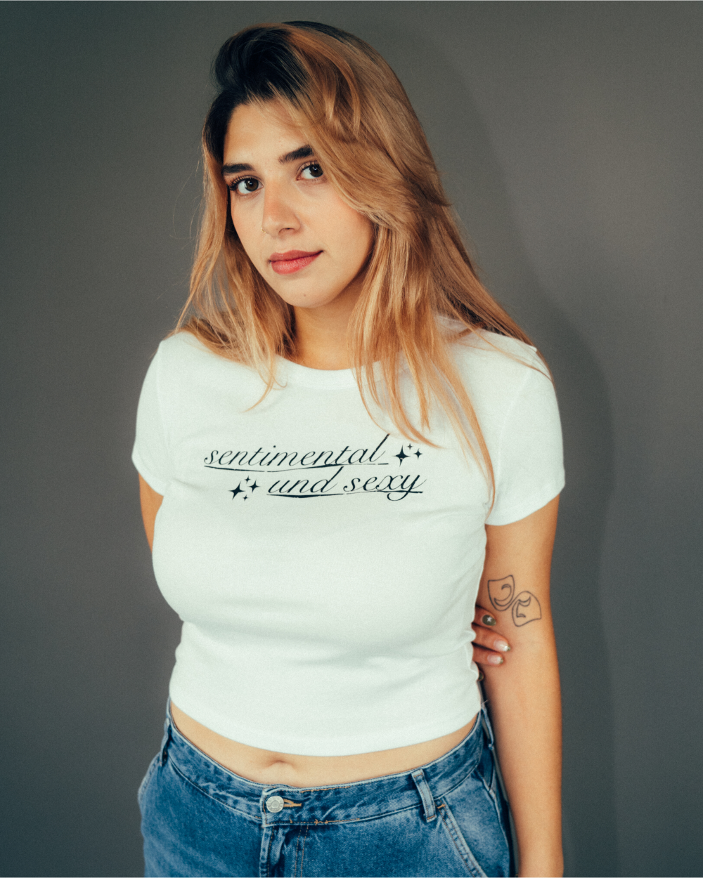 Weißes, tailliertes Crop-T-Shirt mit schwarzem Schriftzug „sentimental and sexy“ in geschwungener Schrift mit kleinen Sternsymbolen. Das Shirt wird mit einer blauen Jeans kombiniert, vor neutralem grauem Hintergrund fotografiert.