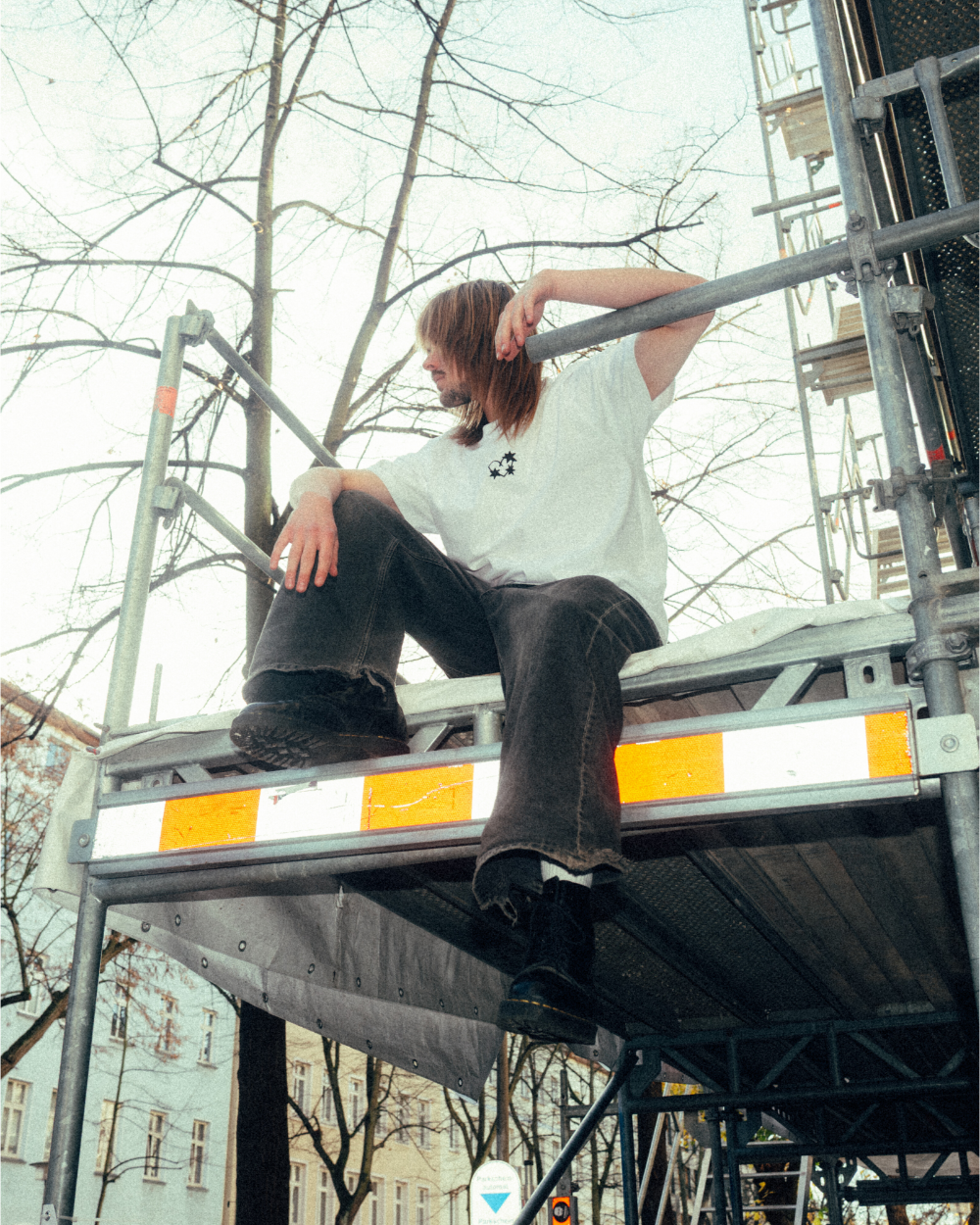 Eine Person sitzt auf einem Baugerüst und trägt ein weißes T-Shirt mit kleinem schwarzen Frontprint auf der linken Brust. Das Motiv zeigt drei miteinander verbundene Dornenherzen. Das Outfit wird mit einer dunkelgrauen Jeans und schwarzen Schuhen kombiniert. Die Szene spielt im Freien, umgeben von kahlen Bäumen und Gebäuden, mit urbanem Charakter und weichem Tageslicht.