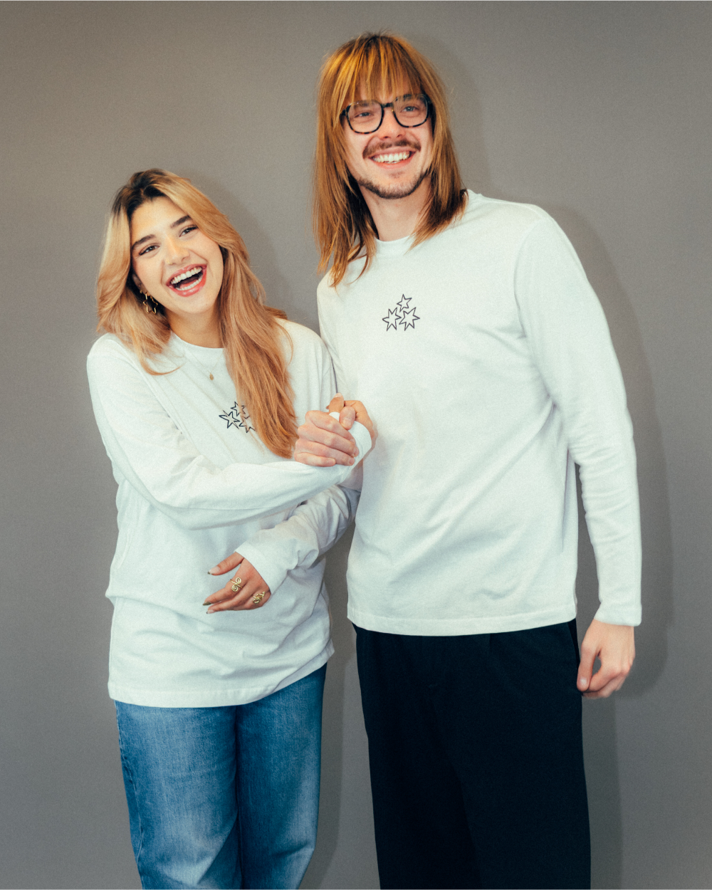 Zwei Personen tragen weiße Longsleeves mit identischem Frontdesign. Auf der linken Brustseite ist eine schwarze Stickerei aus drei sternförmigen Elementen zu sehen, die ein Herzmotiv bilden. Die Shirts bestehen aus weichem Baumwollstoff, haben lange Ärmel und eine locker geschnittene Passform mit Rundhalsausschnitt.