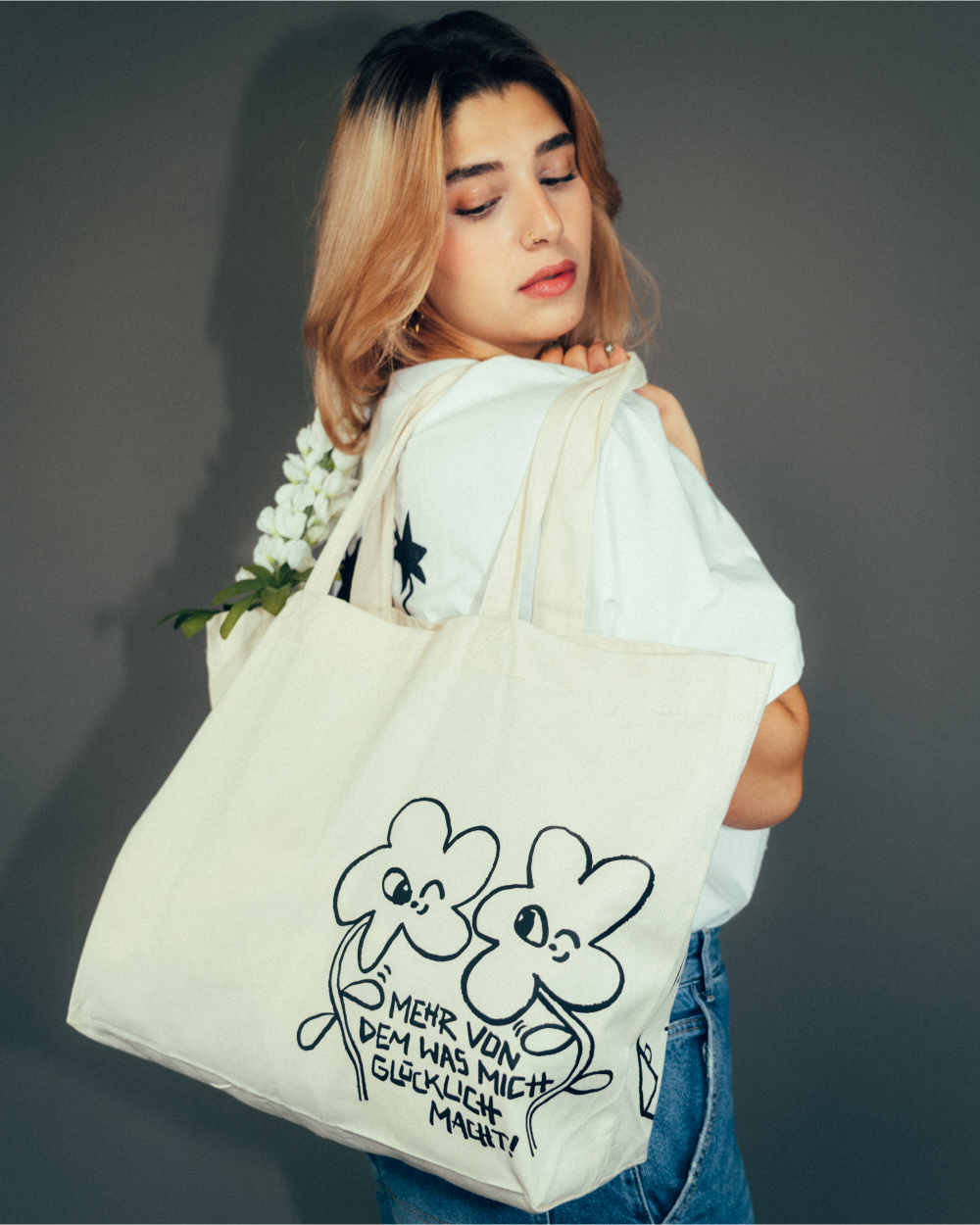 Beiger Baumwoll-Shopper mit schwarzem Print von zwei fröhlich illustrierten vierblättrigen Kleeblättern. Unter den Figuren steht der handgeschriebene Schriftzug „Mehr von dem, was mich glücklich macht!“. Die Tasche wird über der Schulter getragen und mit weißen Blüten dekoriert – verspieltes, positives Accessoire aus Stoff für den Alltag.