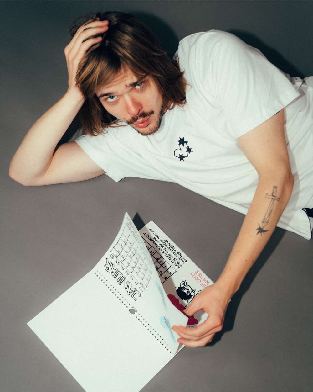 Person liegt auf dem Boden und blättert in einem aufgeklappten Kalender. Sichtbar ist die handgezeichnete Januar-Seite mit einem Monatsraster und illustrativem Text. Der Kalender zeigt den typischen verspielten Stil mit schwarz-weißer Liniengrafik und farbigen Akzenten.