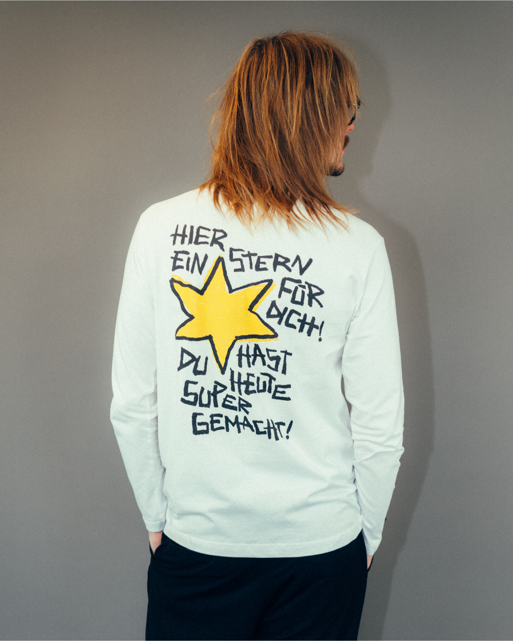 Rückansicht eines weißen Longsleeves aus Baumwolle mit großem, handschriftlich gestaltetem Druck. Das Motiv zeigt einen gelben, skizzenhaften Stern mit schwarzer Umrandung. Darüber und darunter steht in schwarzer Schrift: „HIER EIN STERN FÜR DICH! DU HAST HEUTE SUPER GEMACHT!“. Das Shirt hat eine lockere Passform und lange Ärmel.