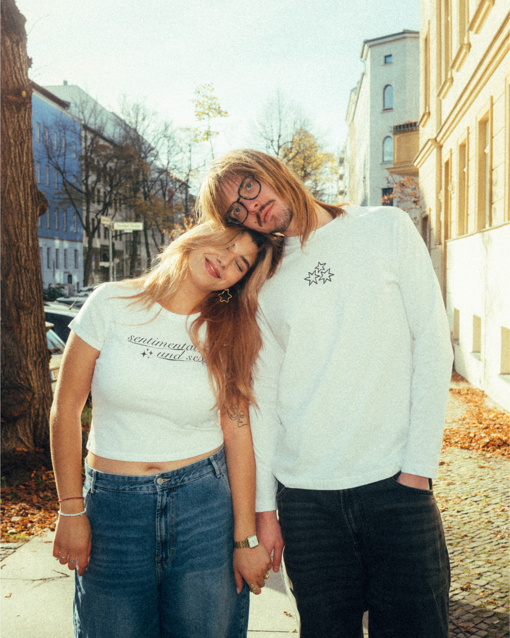Zwei Personen stehen auf einem herbstlichen Gehweg und lehnen sich aneinander. Die linke Person trägt ein weißes, eng geschnittenes Crop-T-Shirt mit dem Schriftzug „sentimental and sexy“ in schwarzer Schreibschrift mit kleinen Sternsymbolen und kombiniert es mit einer blauen Jeans. Die rechte Person trägt ein weißes Langarmshirt mit kleinem schwarzen Sternmotiv auf der Brust und eine dunkle Hose. Beide stehen entspannt in der Sonne zwischen Häuserfassaden und Bäumen.
