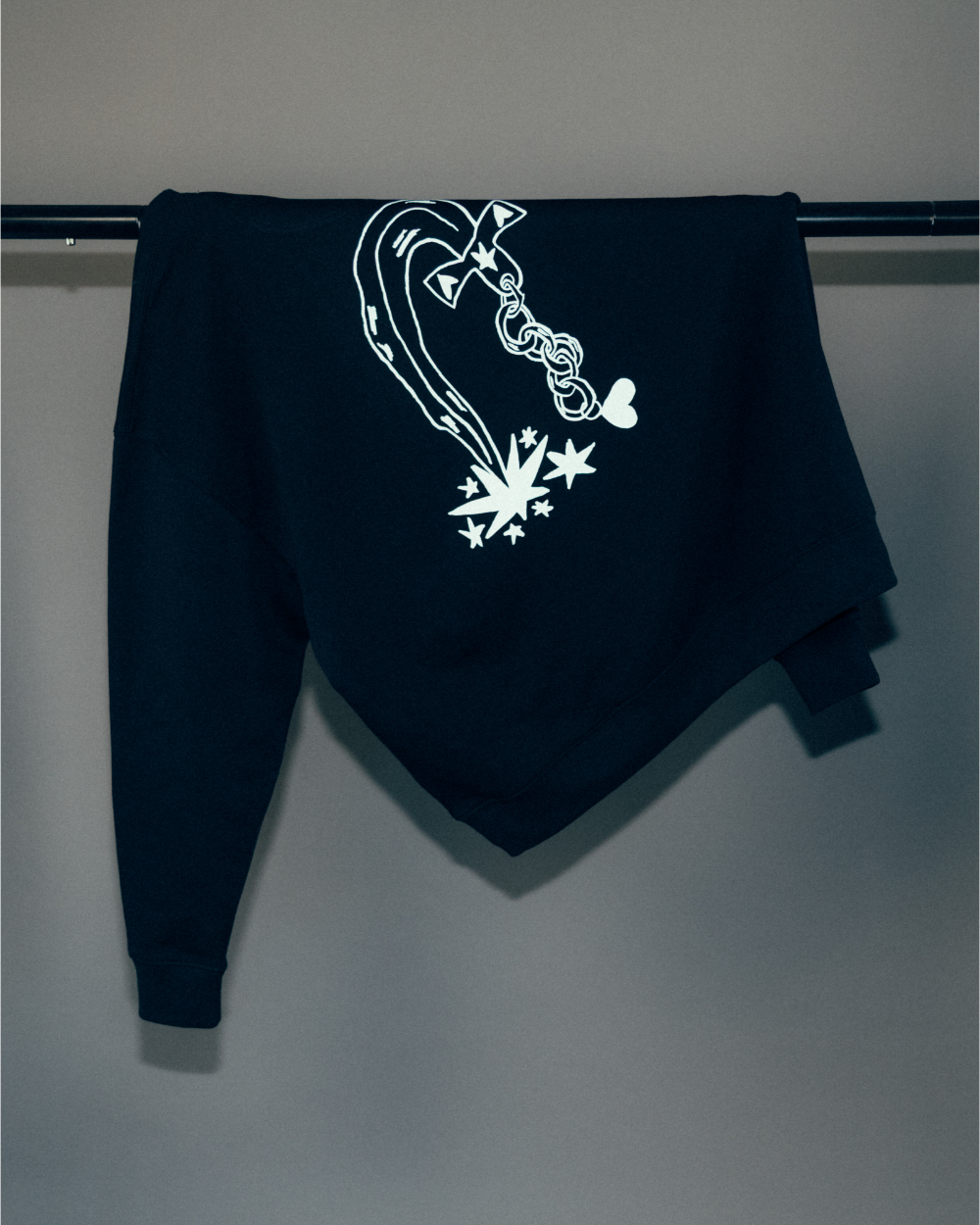 Schwarzer Hoodie, über eine Kleiderstange gehängt, mit großem weißen Rückendruck. Das Motiv zeigt eine stilisierte Kette mit Schleifenform, die in grafisch gestaltete Sterne und Linien übergeht. Der Druck ist mittig auf dem Rücken platziert, die Ärmel enden in elastischen Bündchen.