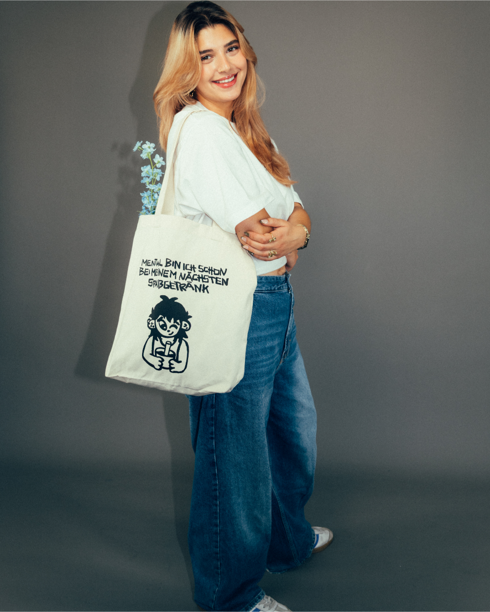 Person steht seitlich und trägt eine beigefarbene Stofftasche mit schwarzem Printmotiv. Der Aufdruck zeigt eine gezeichnete Figur mit Becher und den Schriftzug „MENTAL BIN ICH SCHON BEI MEINEM NÄCHSTEN SPASSGETRÄNK“. Aus der Tasche ragen hellblaue Blumen. Die Person trägt ein helles T-Shirt, eine weite Jeans und helle Sneaker.