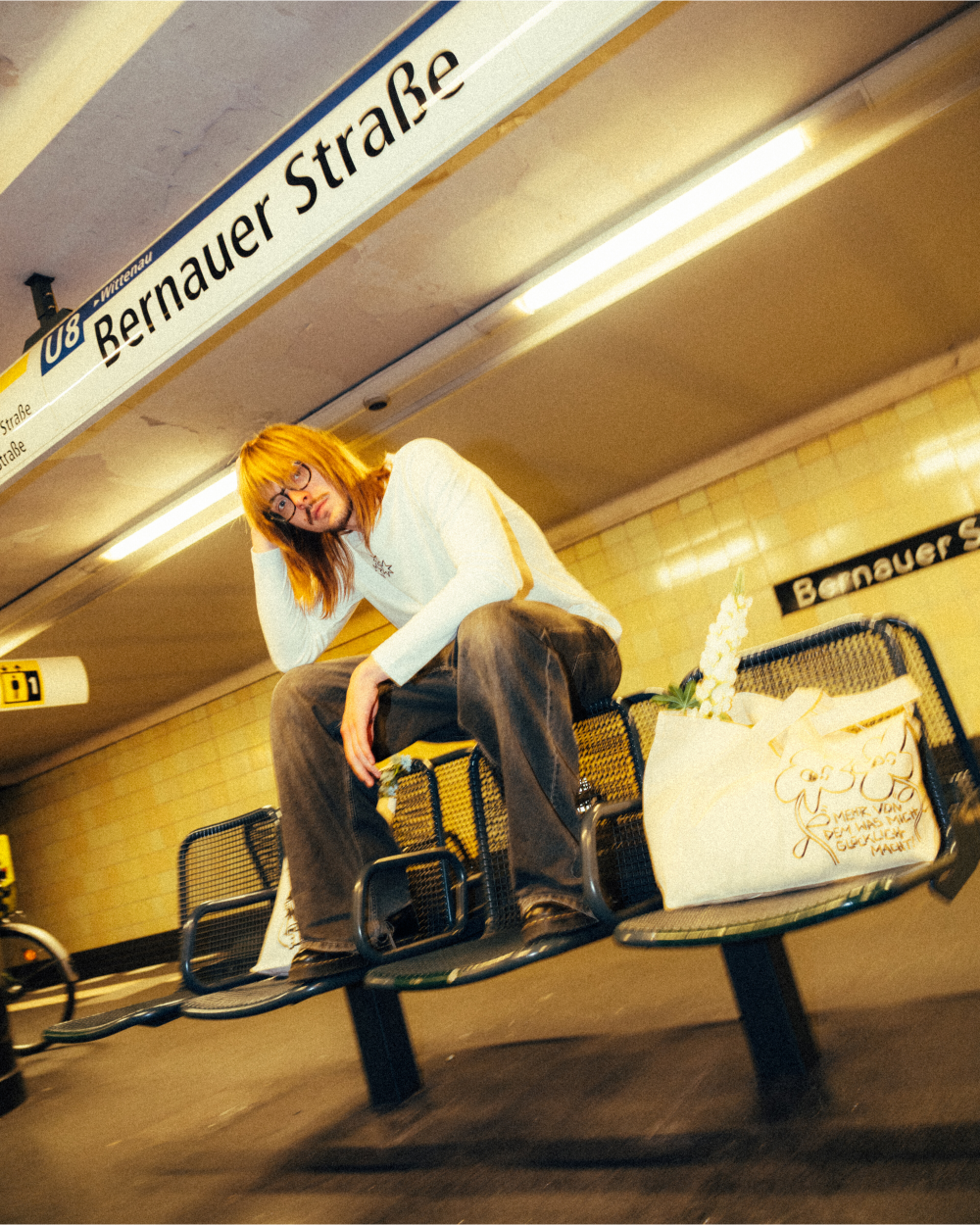 Beiger Stoff-Shopper aus Baumwolle mit schwarzem Print von zwei illustrierten Kleeblättern und dem Schriftzug „Mehr von dem, was mich glücklich macht!“. Die Tasche steht auf einer Sitzbank in einem U-Bahnhof, gefüllt mit weißen Blüten. Alltagstaugliche Stofftasche mit humorvollem Design und positiver Botschaft.