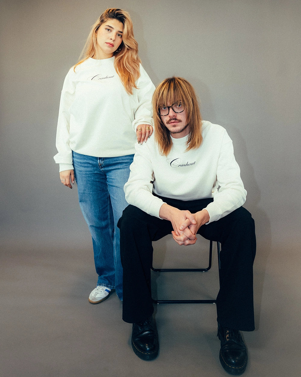 Cremeweißer Sweatshirt mit schwarzem, geschwungenem Schriftzug „Crashout“ mittig auf der Brust. Der Pullover besteht aus weichem Baumwollstoff und hat einen Rundhalsausschnitt sowie gerippte Bündchen an Ärmeln und Saum. Locker geschnittenes Modell mit klassischer Passform, hier im Paar-Look getragen.
