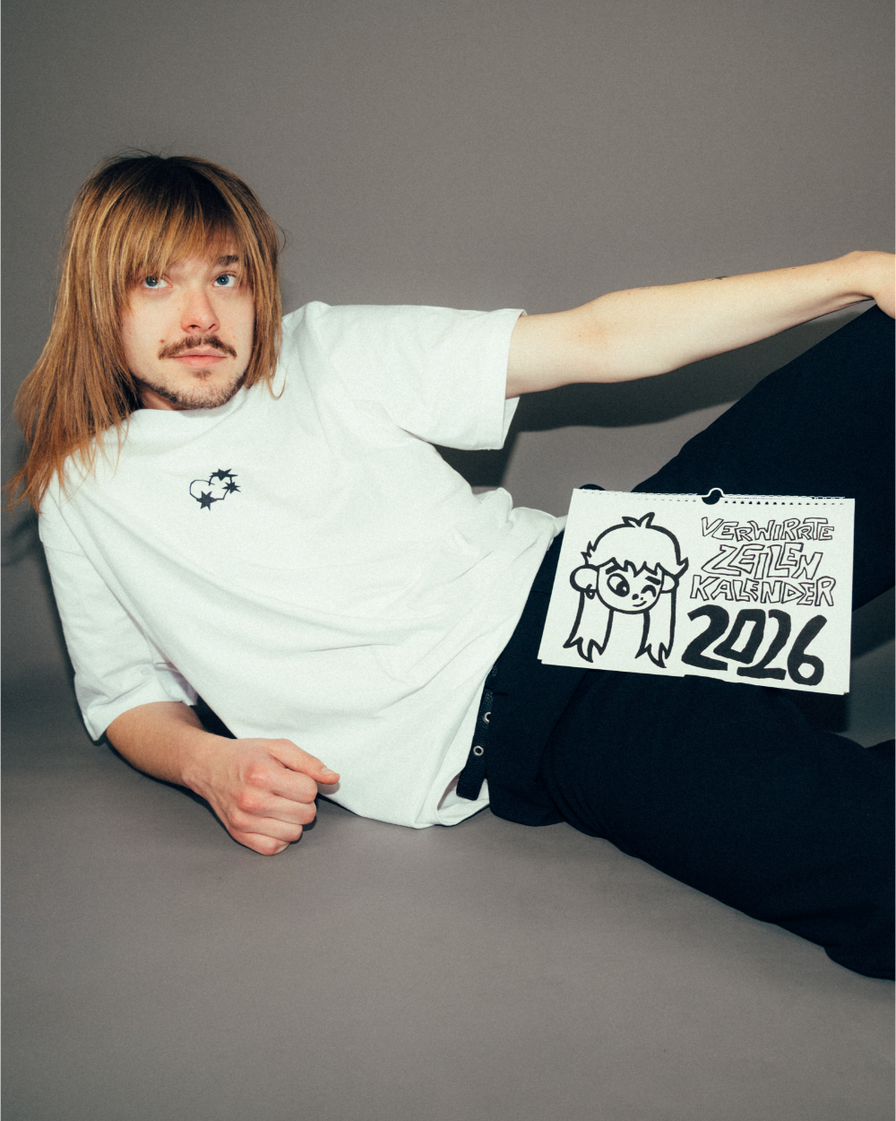 Person liegt in entspannter Pose auf grauem Untergrund und hält den „Verwirrte Zeilen Kalender 2026“ in der Hand. Der Kalender zeigt ein handgezeichnetes Titelmotiv mit einer Comicfigur und typografischem Schriftzug in schwarzer Markeroptik.