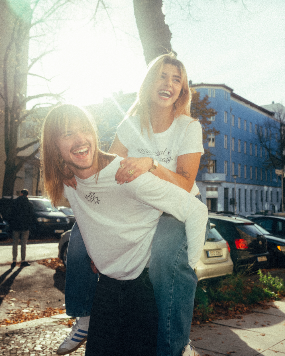 Zwei Personen im Sonnenlicht auf einer herbstlichen Straße; die vordere trägt ein weißes Langarmshirt mit kleinem schwarzen Sternmotiv auf der Brust, die hintere ein weißes Crop-T-Shirt mit dem Schriftzug „sentimental and sexy“ in schwarzer Schreibschrift. Das Shirt wird mit einer blauen Jeans kombiniert. Beide lachen und wirken ausgelassen, während die hintere Person auf dem Rücken der vorderen sitzt.
