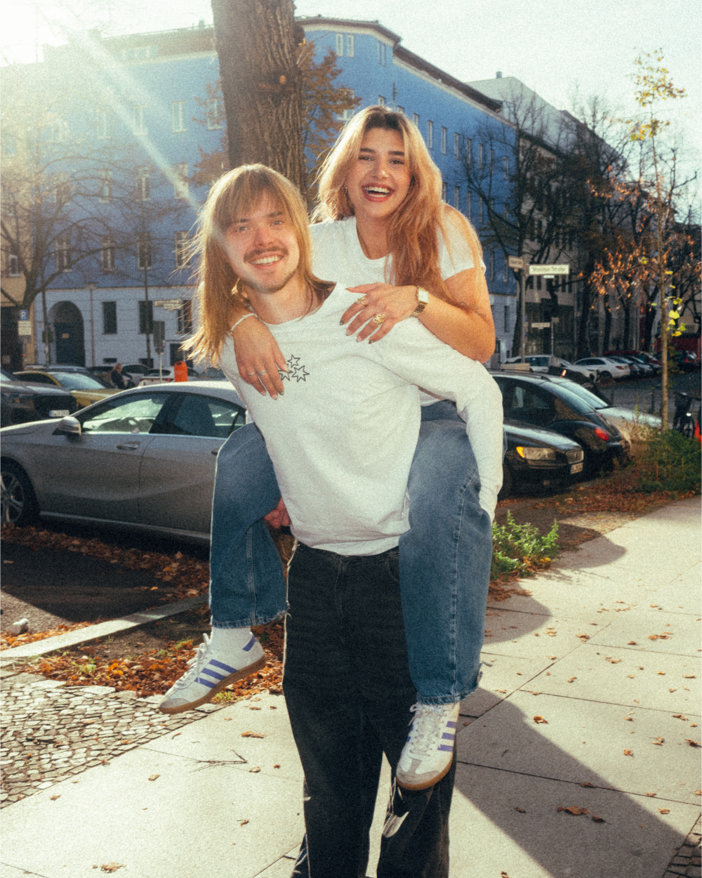 Zwei Personen tragen weiße Oberteile aus Baumwolle in urbaner Umgebung. Das vordere Longsleeve zeigt auf der Brust ein kleines, schwarzes Herzmotiv aus drei sternförmigen Elementen. Das zweite Kleidungsstück ist ein weißes T-Shirt mit schmalem Schnitt und Rundhalsausschnitt. Beide Produkte haben eine legere, alltagstaugliche Passform.