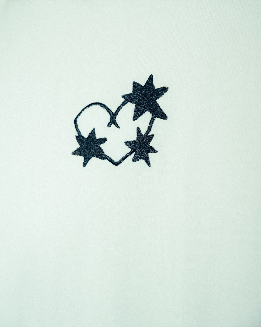 Detailaufnahme eines weißen T-Shirts mit gesticktem schwarzen Frontmotiv. Das Design zeigt drei sternförmige Dornen, die in Form eines Herzens miteinander verbunden sind. Die Stickerei befindet sich auf der linken Brustseite des Shirts.