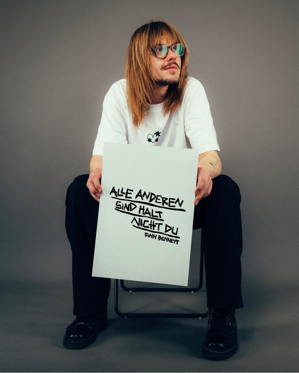 Person sitzt lässig auf einem Stuhl in neutraler Studio-Umgebung und hält ein Blatt mit handschriftlichem Spruch hoch. Darauf steht in schwarzer Markeroptik: „ALLE ANDEREN SIND HALT NICHT DU“ mit der Signatur „FINN BENNETT“. Die Schrift ist teilweise unterstrichen und unregelmäßig gesetzt. Das weiße T-Shirt der Person trägt einen kleinen Print auf Brusthöhe – der Fokus liegt auf dem humorvoll-romantischen Zitatmotiv.