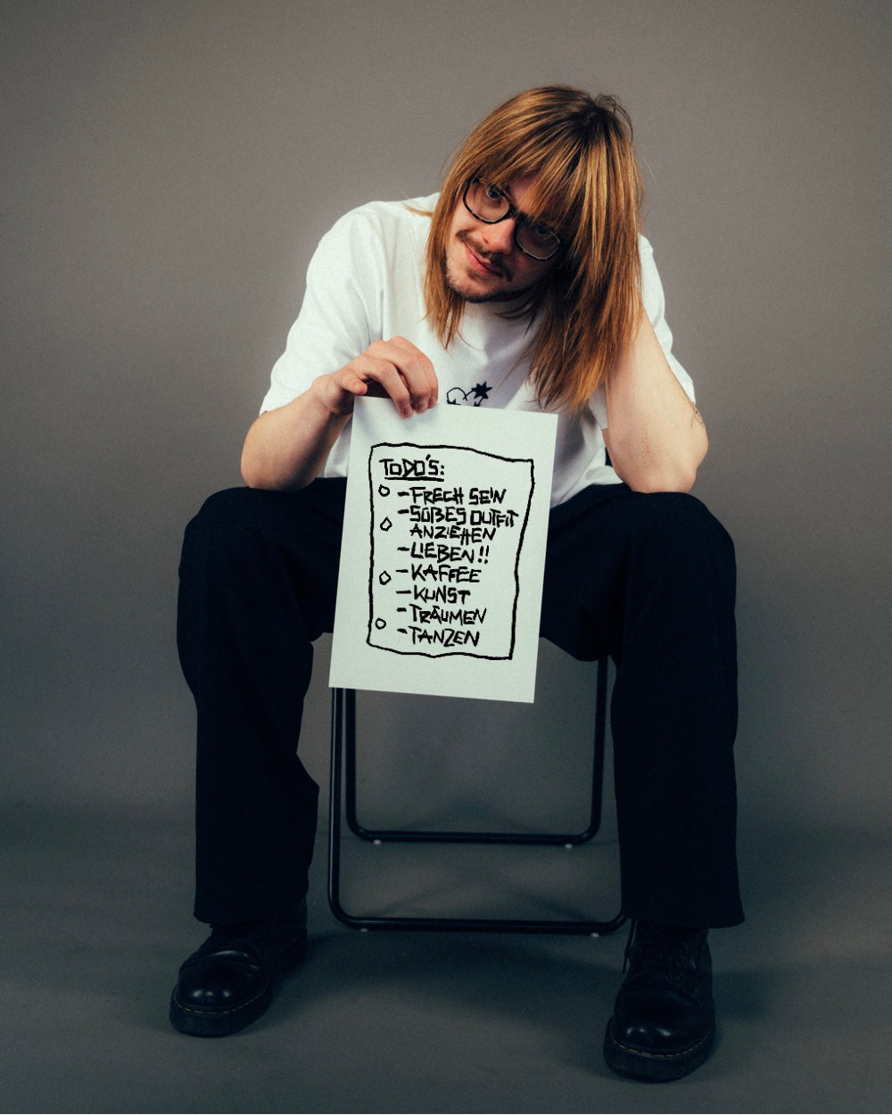 Person in lockerem Outfit mit weißem T-Shirt und dunkler Hose sitzt auf einem Stuhl und hält ein Blatt mit handschriftlicher To-do-Liste hoch. Auf dem Zettel stehen humorvolle Punkte wie „FRECH SEIN“, „SÜßES OUTFIT ANZIEHEN“, „LIEBEN!!“, „KAFFEE“, „KUNST“, „TRÄUMEN“ und „TANZEN“. Der Druck auf dem Shirt ist teilweise sichtbar – der Fokus liegt auf der illustrierten To-do-Liste als verspieltem Printmotiv.