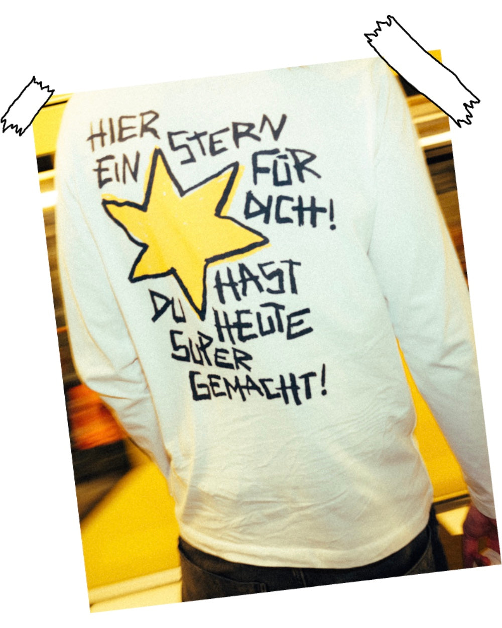 Rückansicht eines weißen Longsleeves mit großem grafischen Rückendruck. Das Design zeigt einen gelben, handgezeichnet wirkenden Stern, umgeben von dem schwarzen Schriftzug „HIER EIN STERN FÜR DICH! DU HAST DU HEUTE SUPER GEMACHT!“. Die Typografie ist in markanter, verspielter Handschrift gestaltet. Das Longsleeve besteht aus weichem Baumwollstoff und wird locker getragen, während im Hintergrund gelbe Farbelemente einer U-Bahn zu sehen sind.