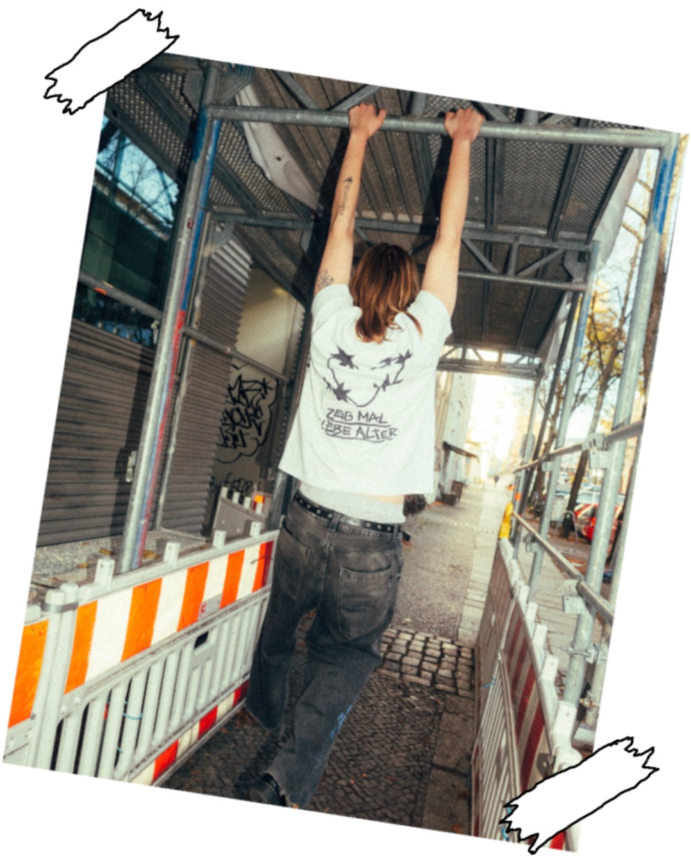 Person trägt ein weißes T-Shirt mit großem schwarzen Rückendruck aus stilisierten Dornenherzen und dem Schriftzug „ZEIG MAL LIEBE ALTER“. Das Shirt wird mit einer dunklen Jeans und einem Gürtel kombiniert. Die Aufnahme zeigt die Rückansicht beim Hochziehen an einem Baugerüst in einer urbanen Straßenszene.