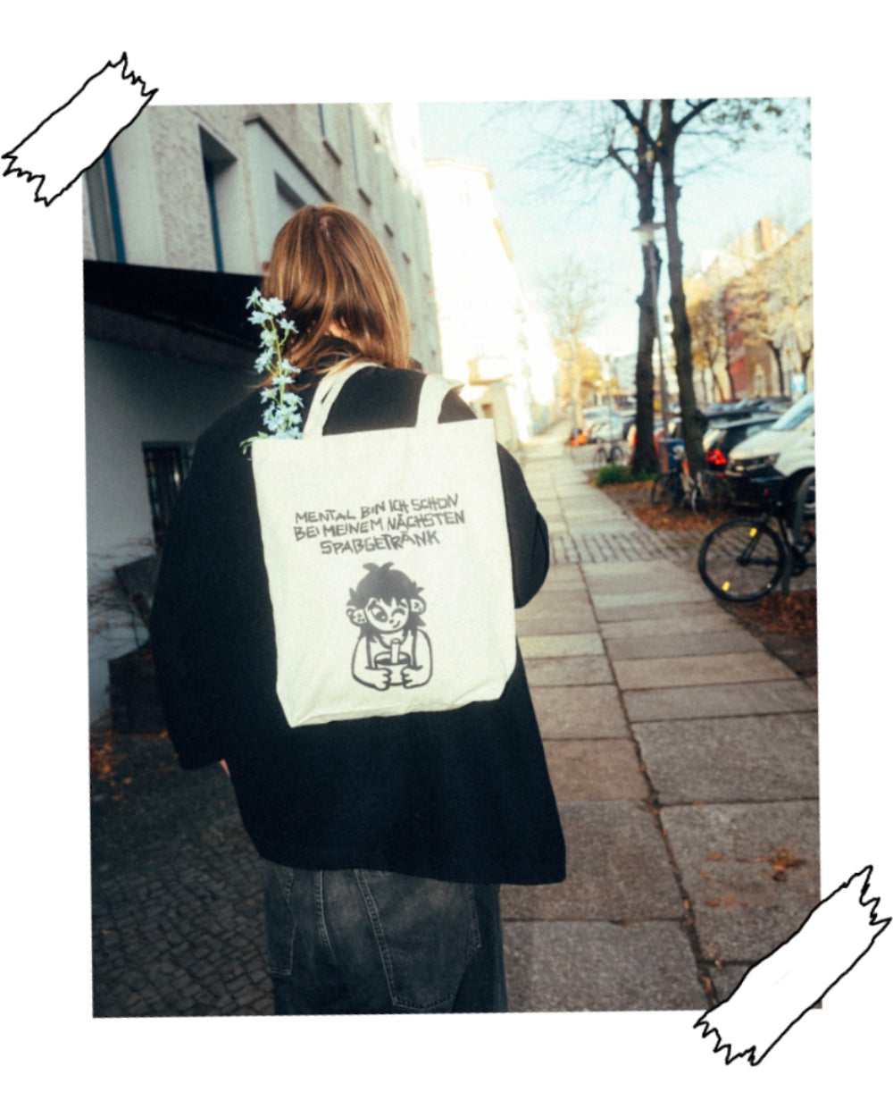 Person geht eine Straße entlang und trägt eine beigefarbene Stofftasche mit schwarzem Aufdruck auf dem Rücken. Das Motiv zeigt eine handgezeichnete Figur mit Becher und den Schriftzug „MENTAL BIN ICH SCHON BEI MEINEM NÄCHSTEN SPASSGETRÄNK“. Aus der Tasche ragen hellblaue Blumen.