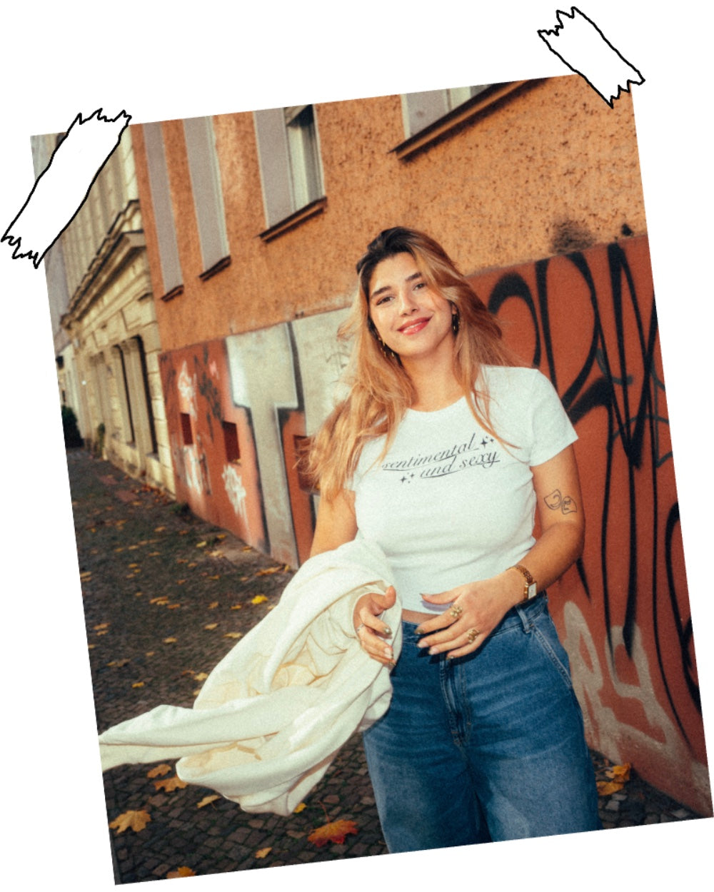 Weiße, kurz geschnittene Babytee mit schwarzem Schriftzug „sentimental and sexy“ auf der Brust. Der Druck ist in geschwungener Schreibschrift mit kleinen Sternsymbolen gestaltet. Die Person trägt dazu eine weite Jeans und goldenen Schmuck, fotografiert in einer herbstlichen Straßenszene mit Graffitiwand im Hintergrund.